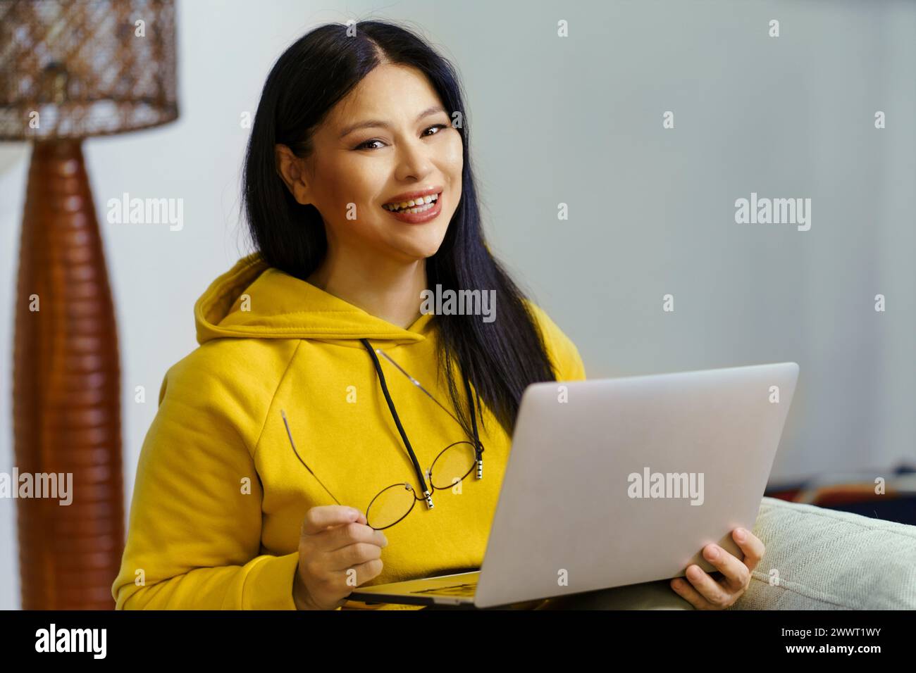 Une femme dans un sweat à capuche jaune sourit et tient un ordinateur portable. Elle porte des lunettes et profite de son temps Banque D'Images