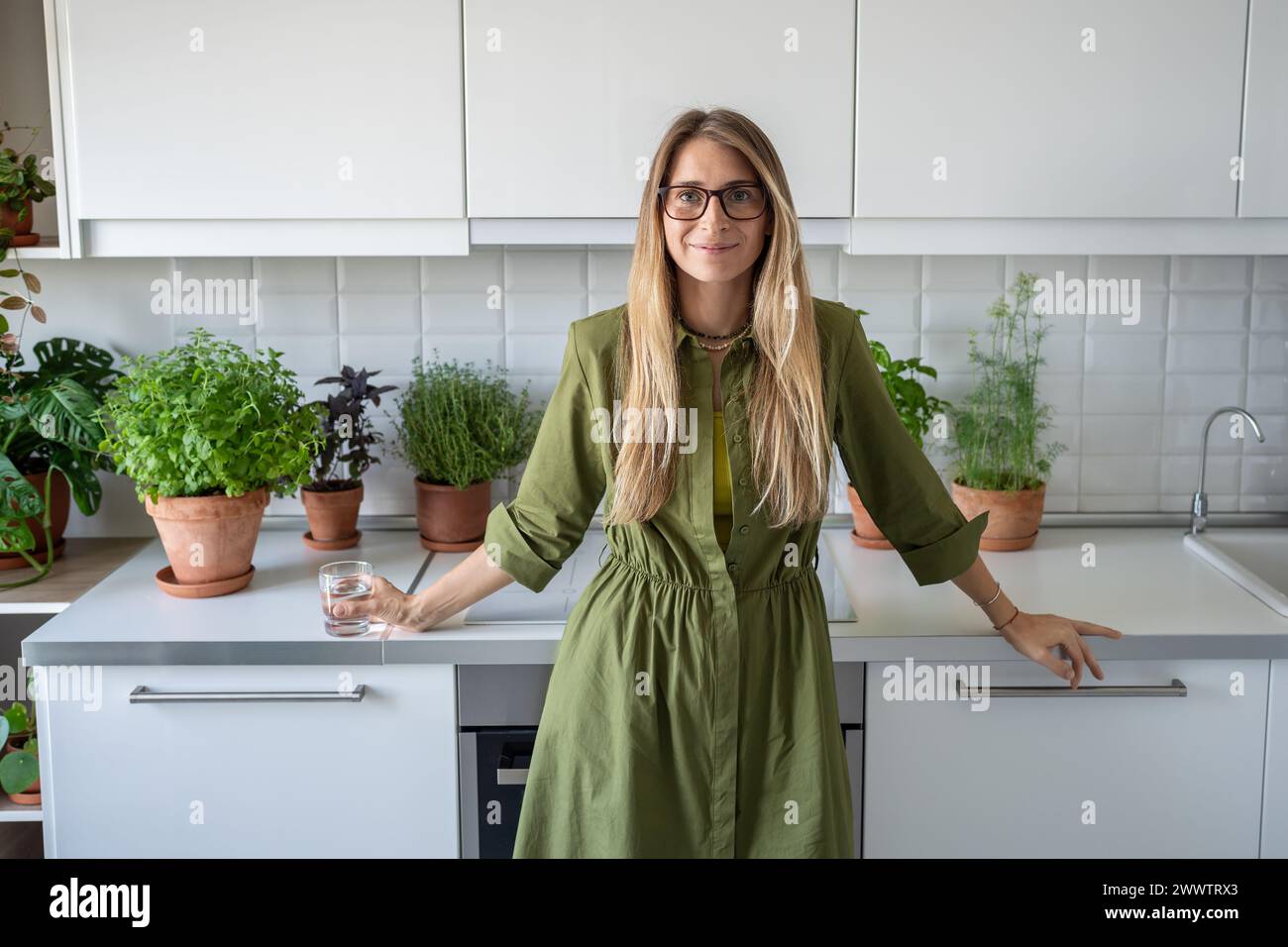 Femme d'âge moyen satisfaite dans des lunettes avec un verre d'eau regardant la caméra debout dans la cuisine. Banque D'Images