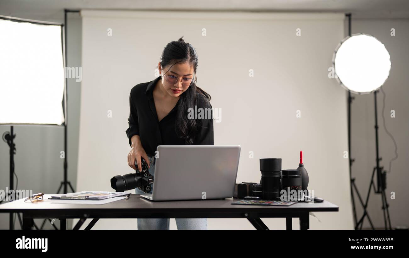 Une photographe asiatique professionnelle et confiante vérifie des images sur un ordinateur portable, travaillant sur une séance photo dans un studio avec un équipement d'éclairage Banque D'Images