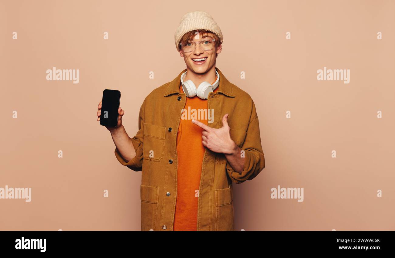 Heureux jeune homme dans des vêtements décontractés se tient dans un studio vibrant. Il tient un téléphone portable, pointant et recommandant une application sur l'écran tactile. Usure g Banque D'Images