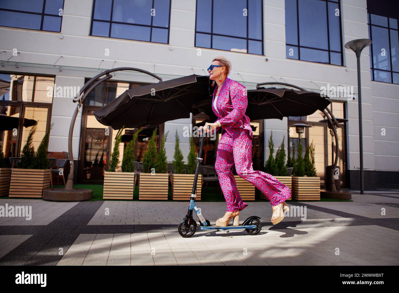 Élégamment habillée femme mature dans un pantalon rose vif et une veste roule autour de la ville sur un scooter, un frien actif, sain et éco Banque D'Images