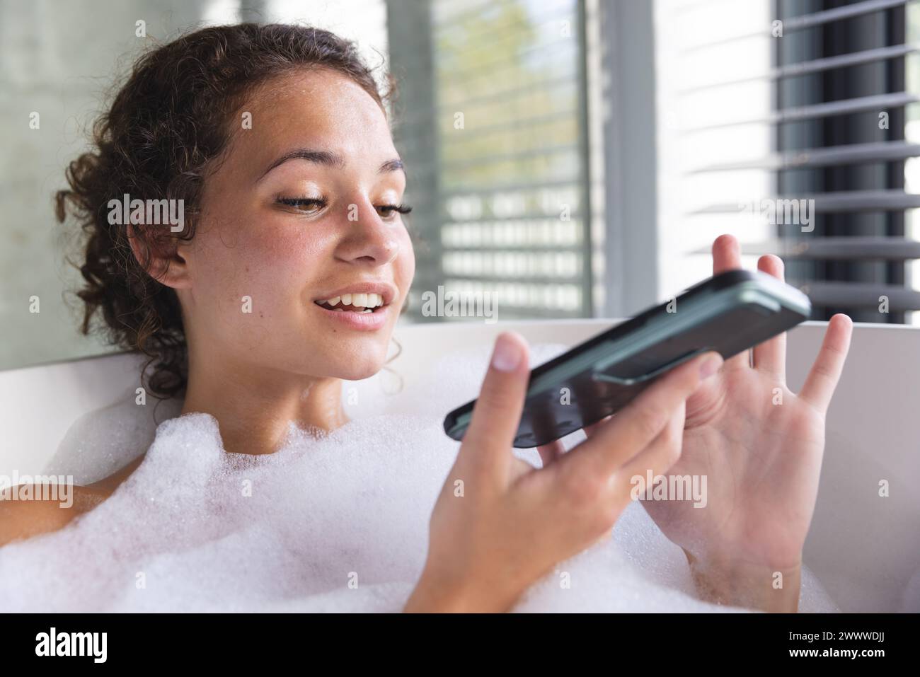 Femme caucasienne se détend dans un bain moussant à la maison, smartphone à la main Banque D'Images