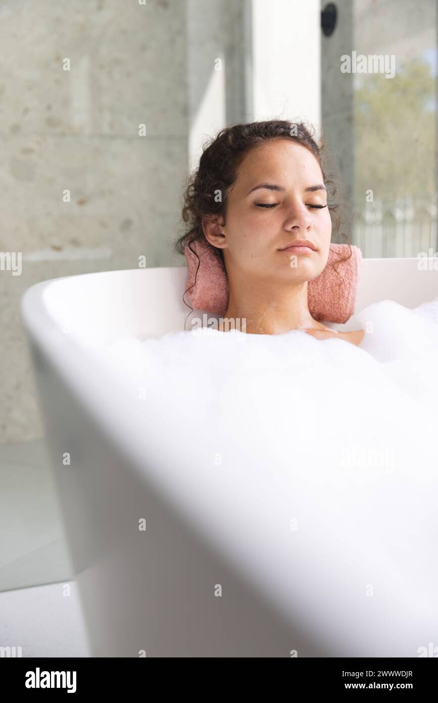 Jeune femme se détend dans un bain moussant à la maison, les yeux fermés dans la sérénité, avec espace copie Banque D'Images