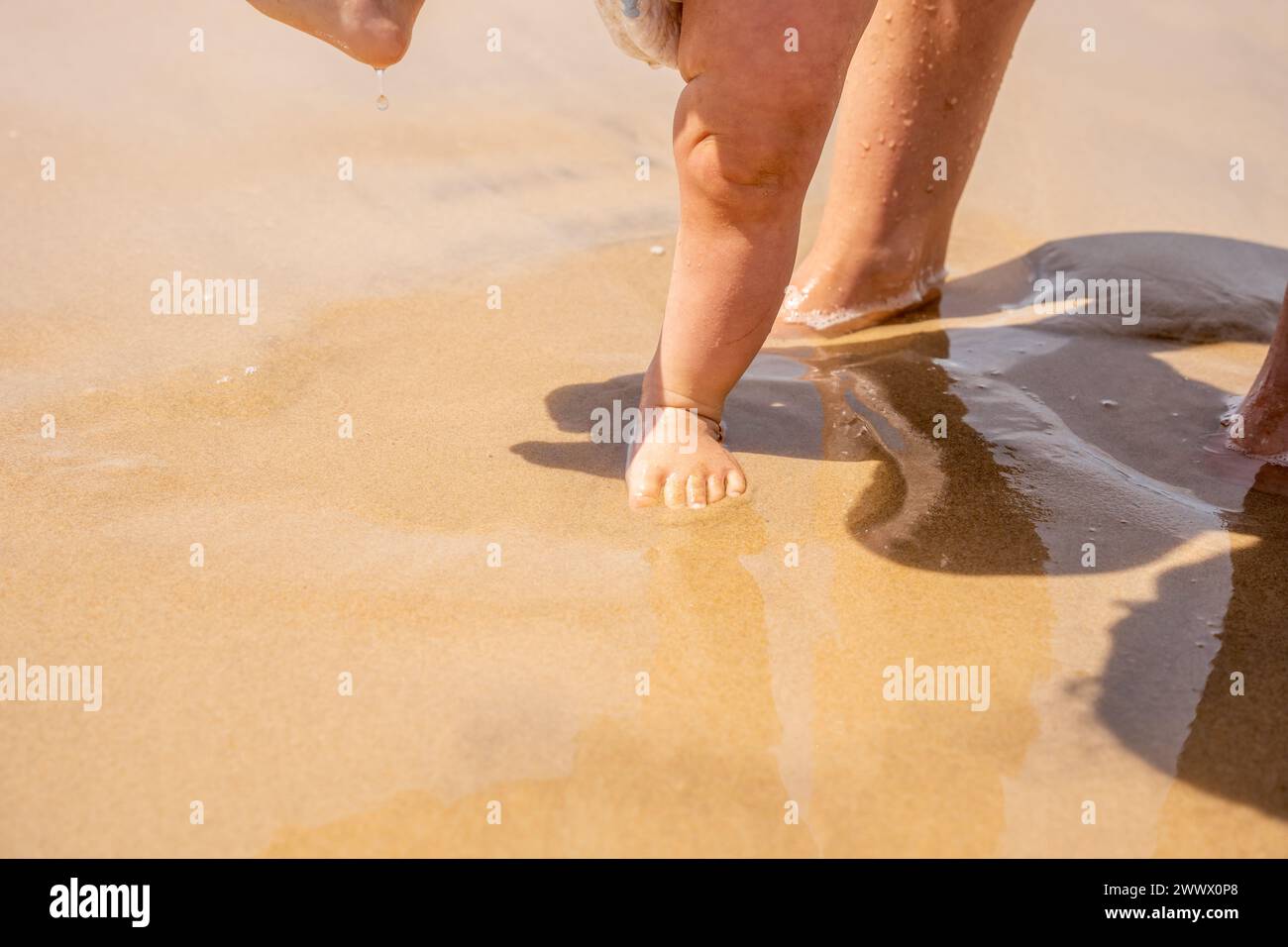 Photographie horizontale des petits pieds d'un bébé marchant sur le sable de la plage pour la première fois, profitant de l'eau et des vagues de la mer. concp Banque D'Images