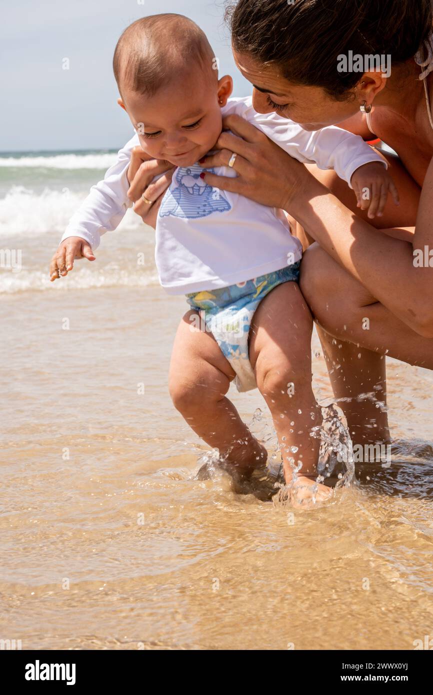 Photographie verticale d'une jeune mère et de son bébé profitant de la plage pour la première fois, jouant dans l'eau et sautant avec les vagues de la mer. Banque D'Images