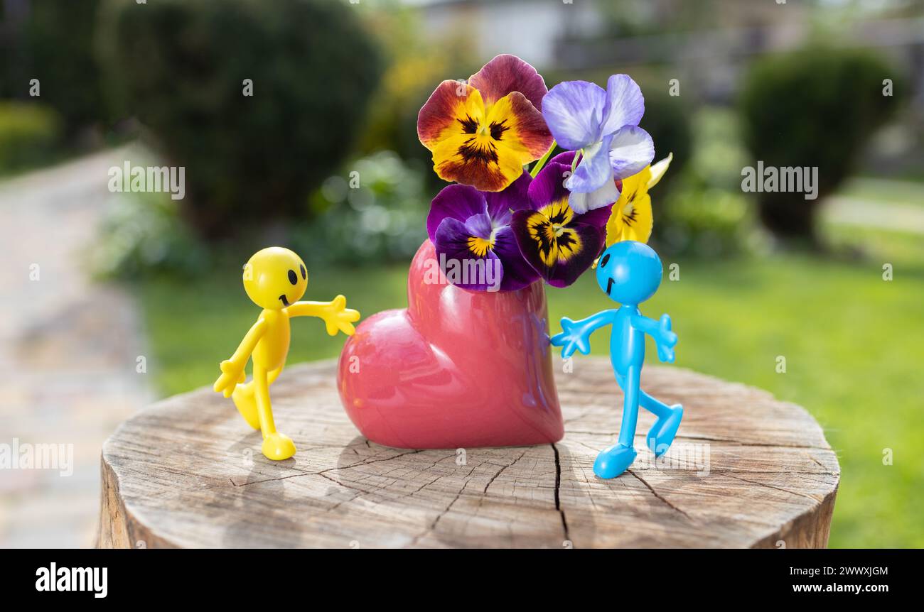 Figurines humaines miniatures en jaune et bleu près d'un vase en forme de coeur avec un bouquet de pansies Banque D'Images