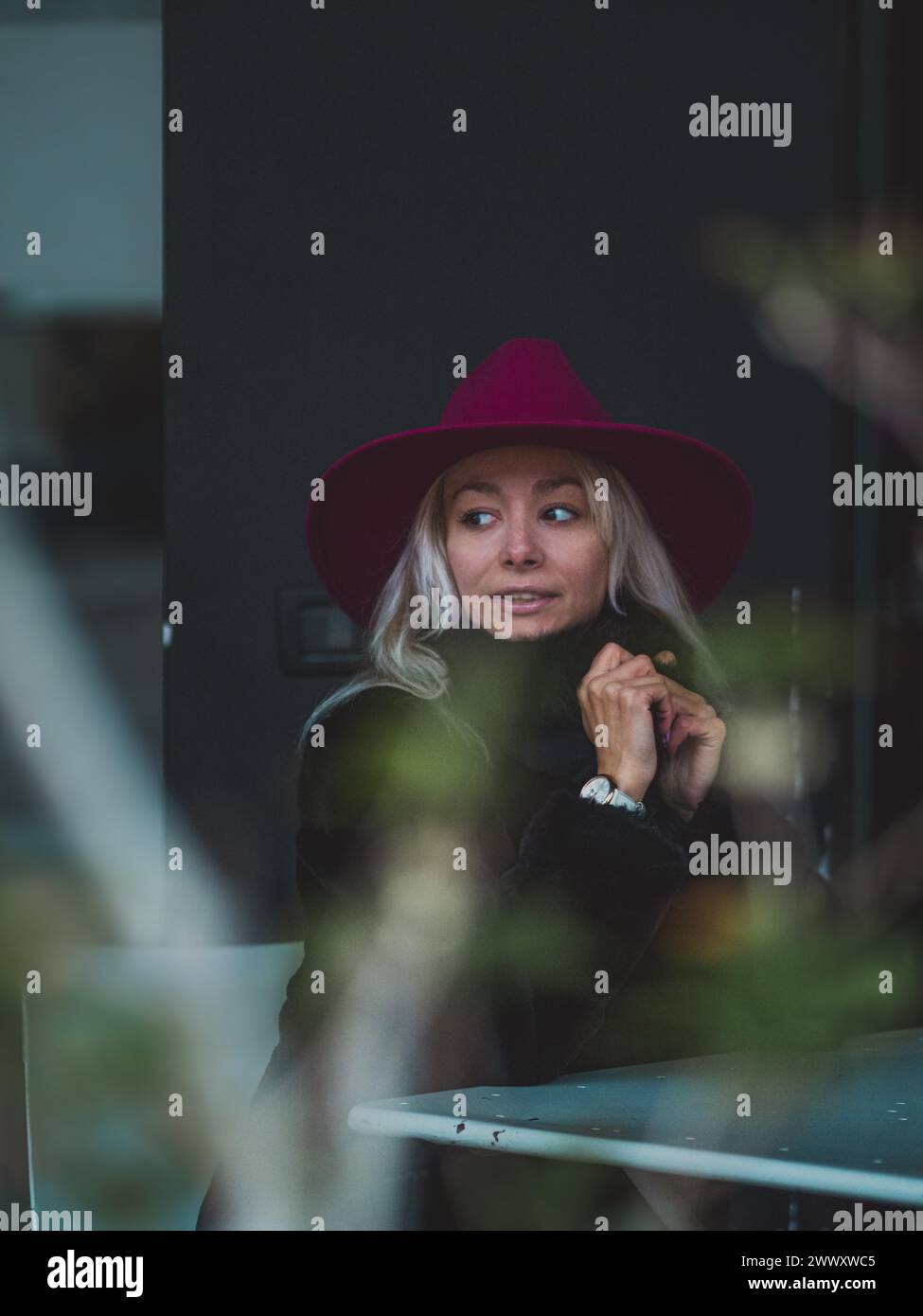 Femme surprise dans un chapeau rouge assis à l'extérieur dans un bar avec la lumière naturelle filtrant à travers la verdure Banque D'Images