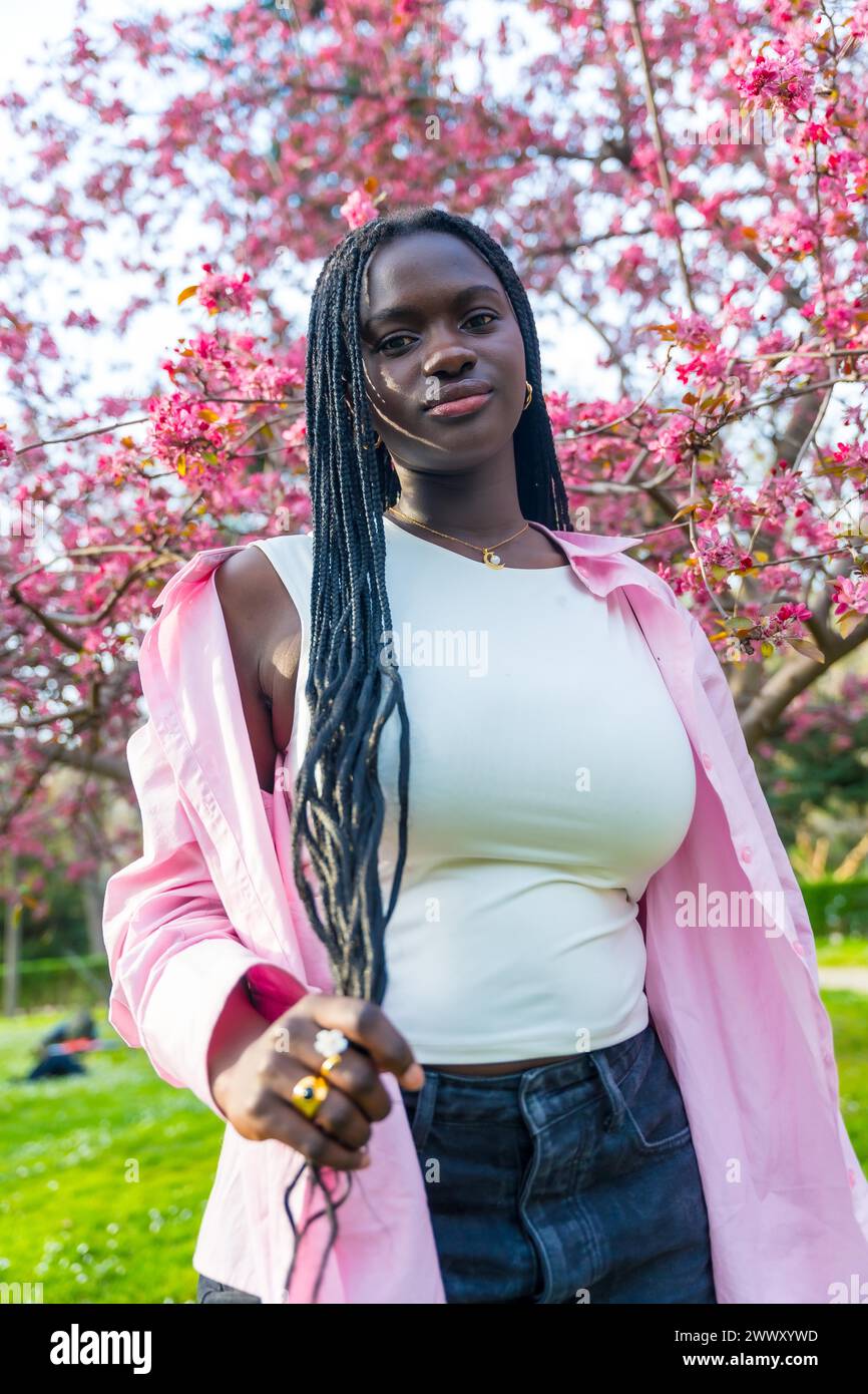 Portrait vertical d'une jeune femme africaine en vêtements décontractés posant à côté d'un arbre rose fleuri Banque D'Images