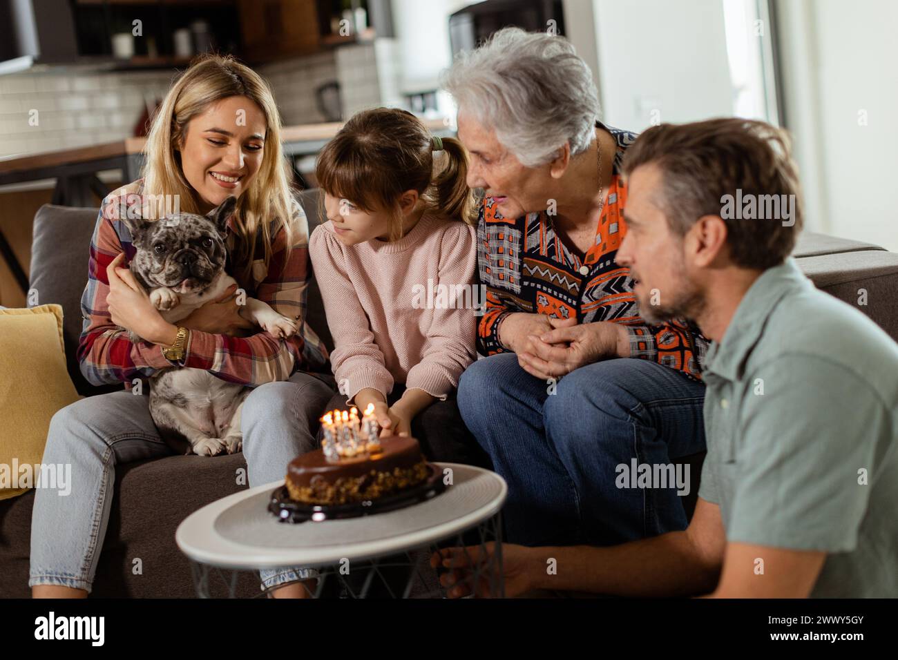 Une scène réconfortante se déroule alors qu'une famille multigénérationnelle se réunit sur un canapé pour présenter un gâteau d'anniversaire à une grand-mère ravie, créant ainsi des souvenirs Banque D'Images