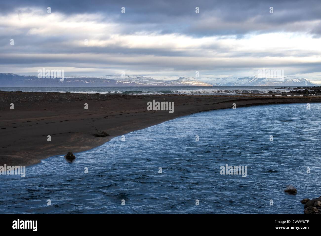 Sable volcanique foncé à côté de la rivière qui coule vers l'océan. Montagnes avec des falaises à l'horizon, avec des sommets de neige. Région de Isafjordur, Westfjords, Icela Banque D'Images