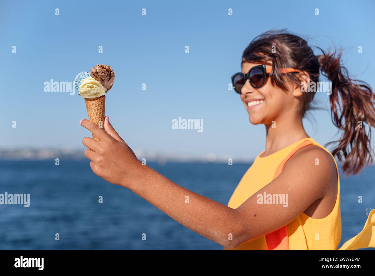 Fille heureuse et souriante tenant le cône de crème glacée avec des boules colorées de crème glacée. Littoral marin ensoleillé à l'arrière-plan. Banque D'Images