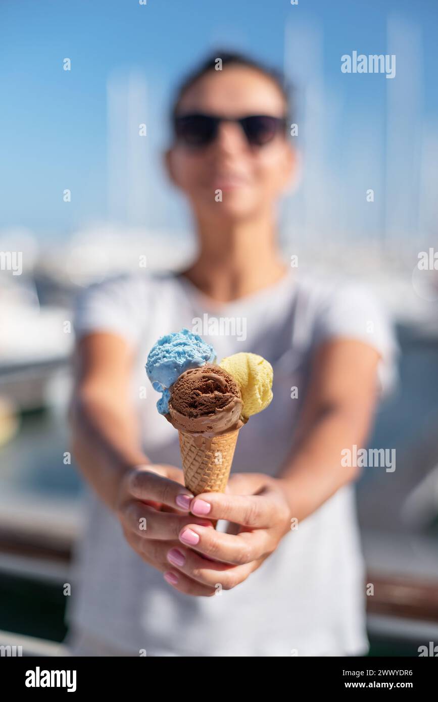 Femme heureuse et souriante tenant un cône de crème glacée avec des boules de crème glacée colorées. Côte ensoleillée de la mer en arrière-plan. Banque D'Images