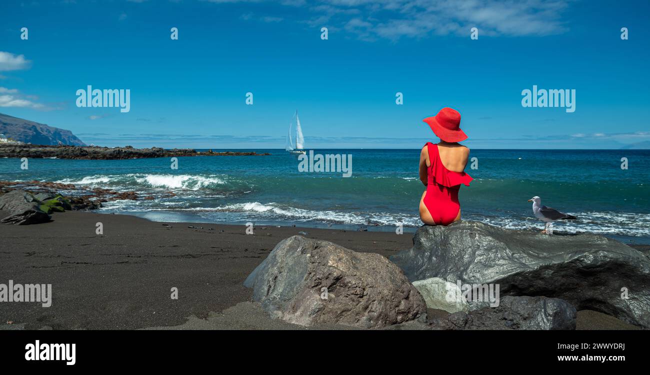 Femme en maillot de bain rouge assis sur le rocher dans la plage de sable de l'océan noir. Eau de l'océan calme et ciel bleu pur à l'arrière-plan. Banque D'Images