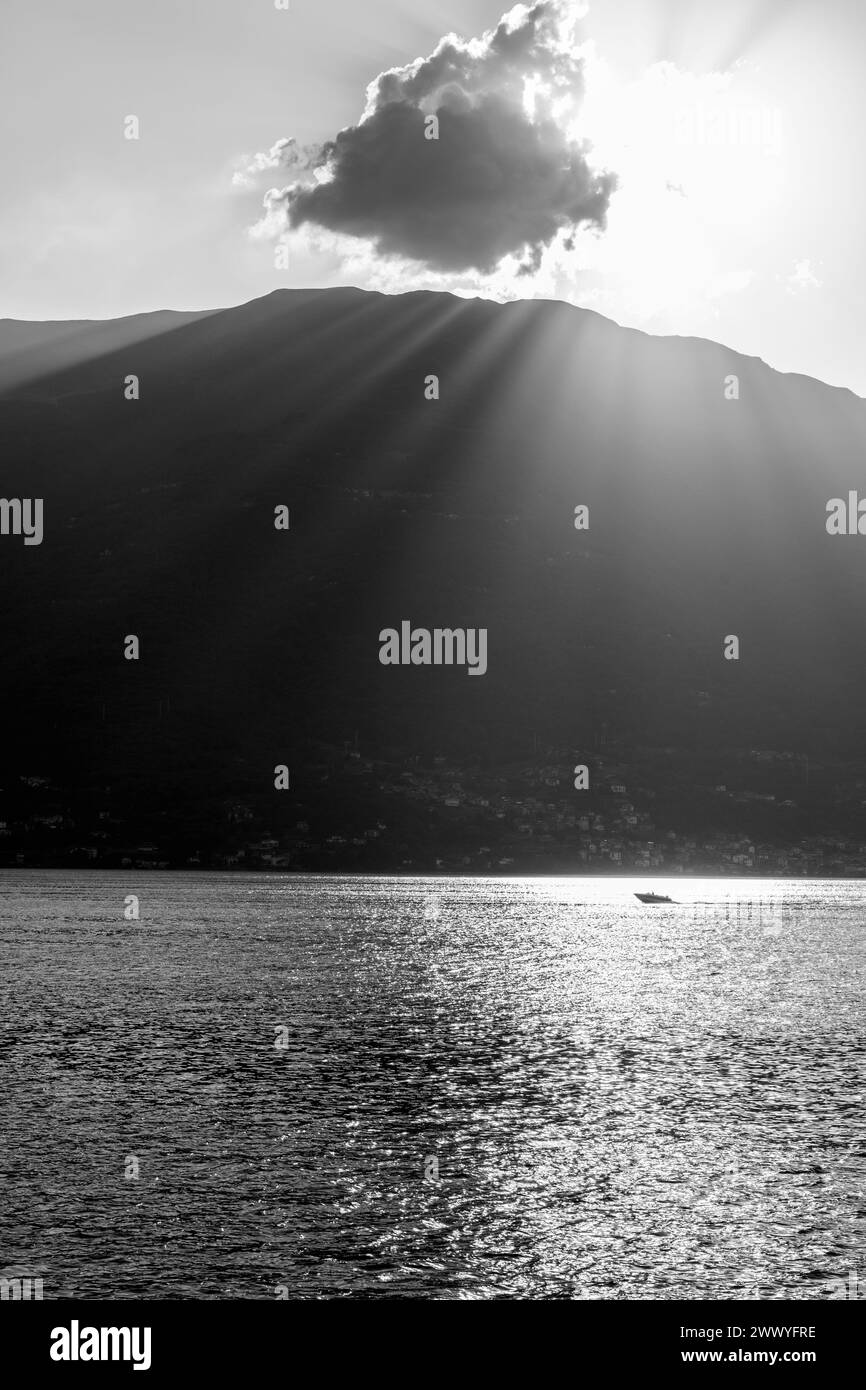 Une scène tranquille capturant la beauté sereine d'un lac de montagne au coucher du soleil. Les rayons du soleil traversent un nuage, projetant des faisceaux lumineux et des reflets étincelants sur la surface de l'eau. Un bateau solitaire profite de l'atmosphère paisible, entouré par la silhouette des montagnes. Image en noir et blanc. Banque D'Images