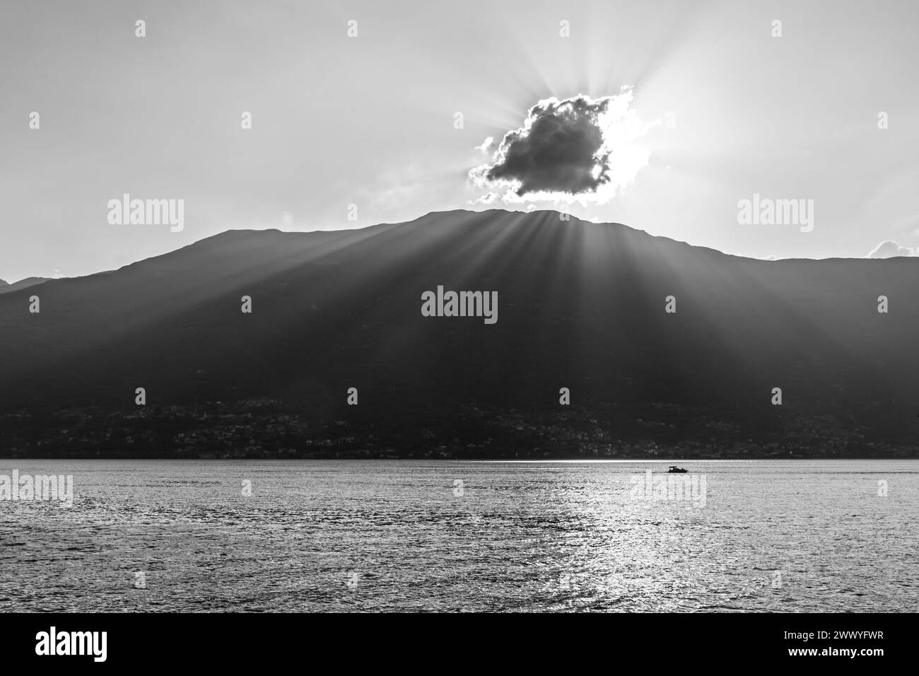 Une scène tranquille capturant la beauté sereine d'un lac de montagne au coucher du soleil. Les rayons du soleil traversent un nuage, projetant des faisceaux lumineux et des reflets étincelants sur la surface de l'eau. Un bateau solitaire profite de l'atmosphère paisible, entouré par la silhouette des montagnes. Image en noir et blanc. Banque D'Images