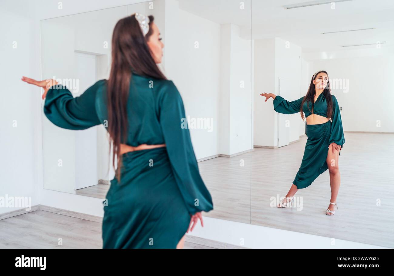 Femme gracieuse habillée en robe de danse latine vert foncé faisant des pas de danse élégants dans la grande salle de couleur blanche avec grand mur miroir. Expressions du peuple du Banque D'Images