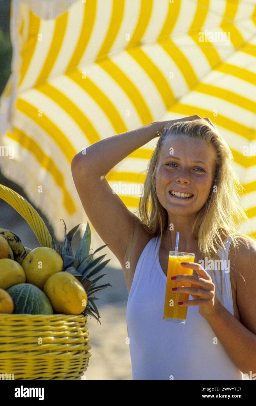 joli cheveux blonds jeune femme attrayante regardant la caméra avant heathy sourire buvant du jus d'orange un parapluie jaune panier de fruits côté sur le fond Banque D'Images