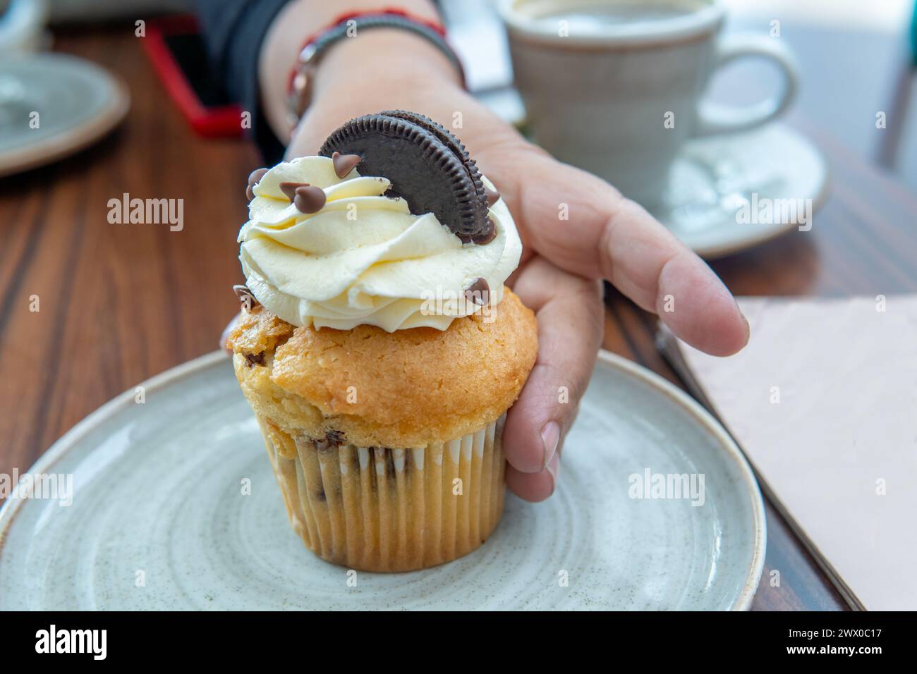 gros plan d'une main prenant un cupcake avec de la crème, des gouttes de chocolat et un cookie sur une table Banque D'Images