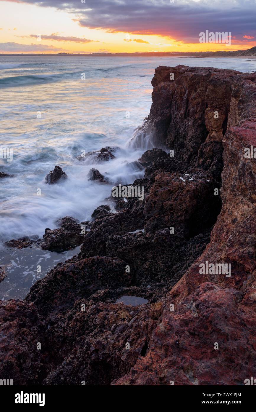 Des vagues s'écrasent sur la base d'une falaise côtière devant un ciel de coucher de soleil à Forrest Caves sur Phillip Island dans le Victoria, Australie. Banque D'Images
