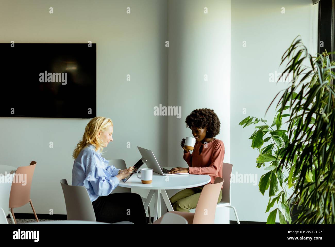 Deux collègues appréciant une conversation joyeuse autour d'un café dans un cadre de bureau moderne et ensoleillé. Banque D'Images