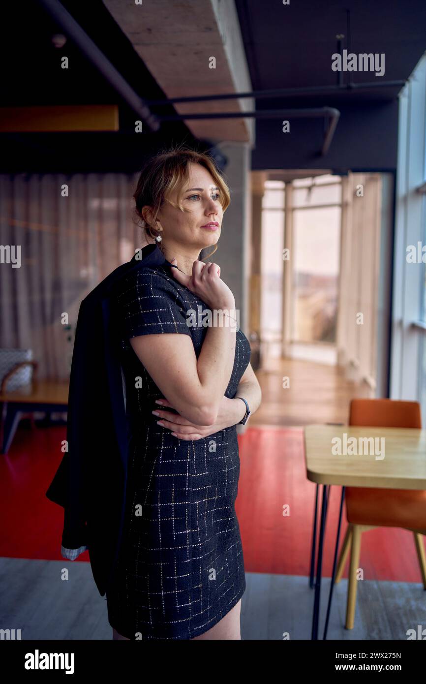une femme attrayante dans un style d'affaires mini robe noire se promène dans un espace de coworking moderne avec des fenêtres panoramiques et un plafond noir Banque D'Images