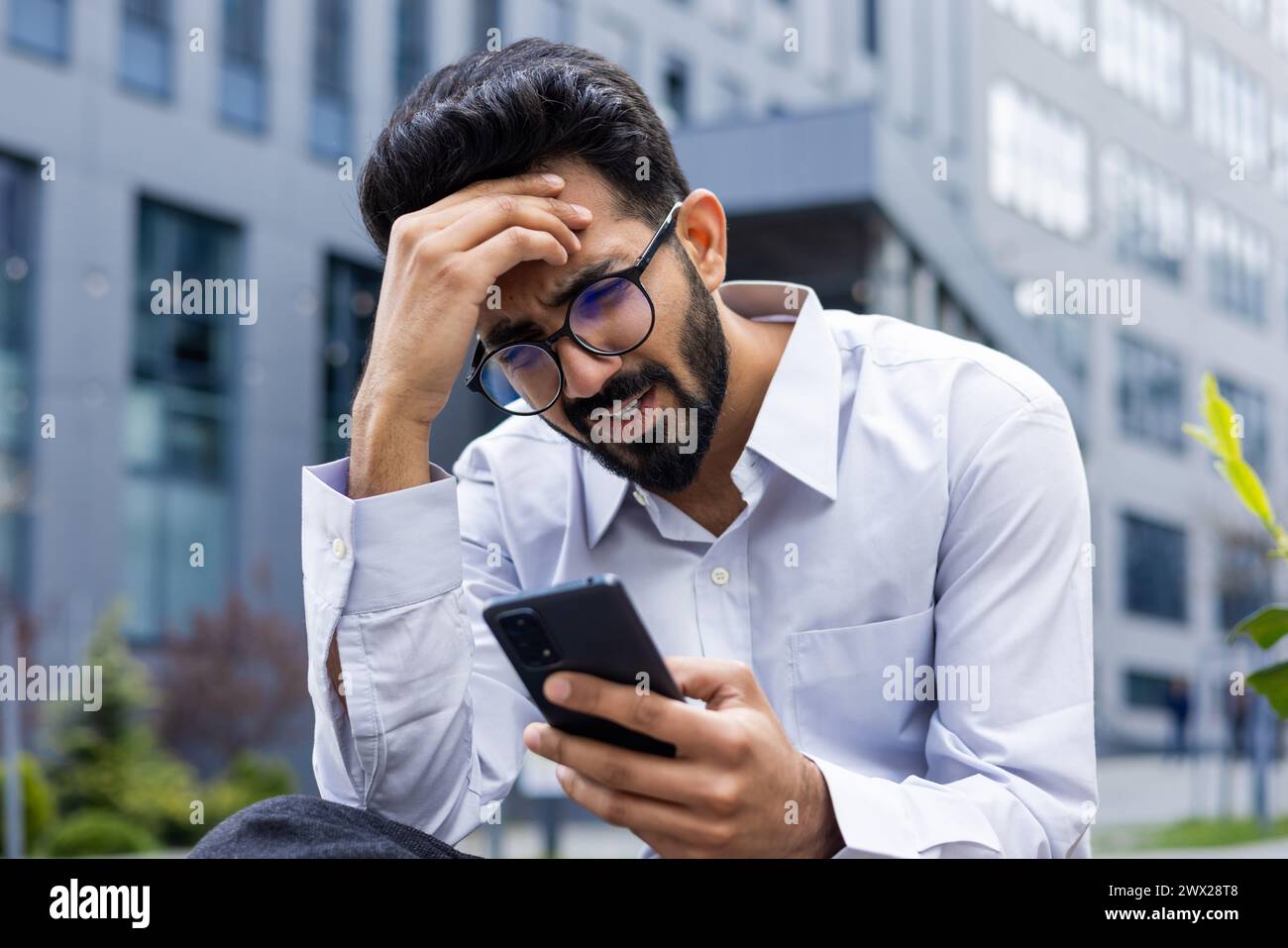 Photo en gros plan d'un jeune homme indien choqué et bouleversé assis à l'extérieur tenant la tête avec la main et regardant l'écran du téléphone après avoir reçu de mauvaises nouvelles. Banque D'Images