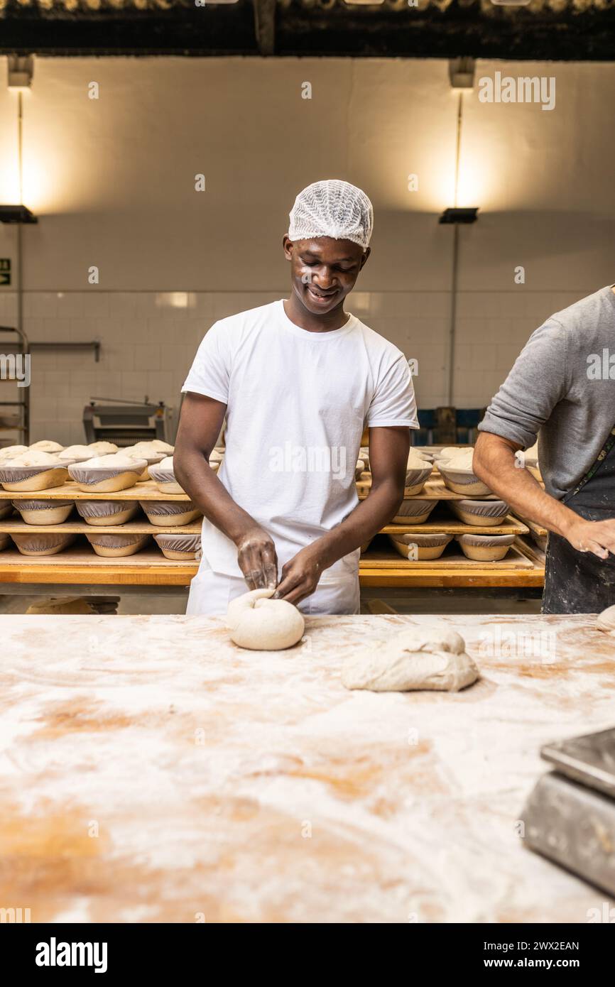 Jeune homme noir souriant travaillant dans une fabrique de pain, apprenant le métier. Banque D'Images