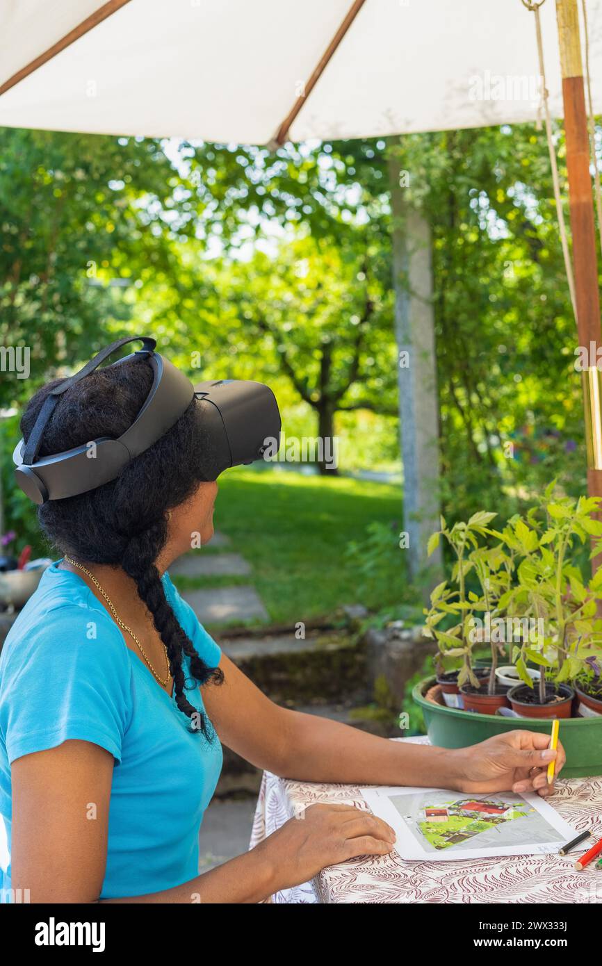 Une femme mixte dans des lunettes VR conçoit un plan de jardin avec des crayons de couleur à une table. Banque D'Images