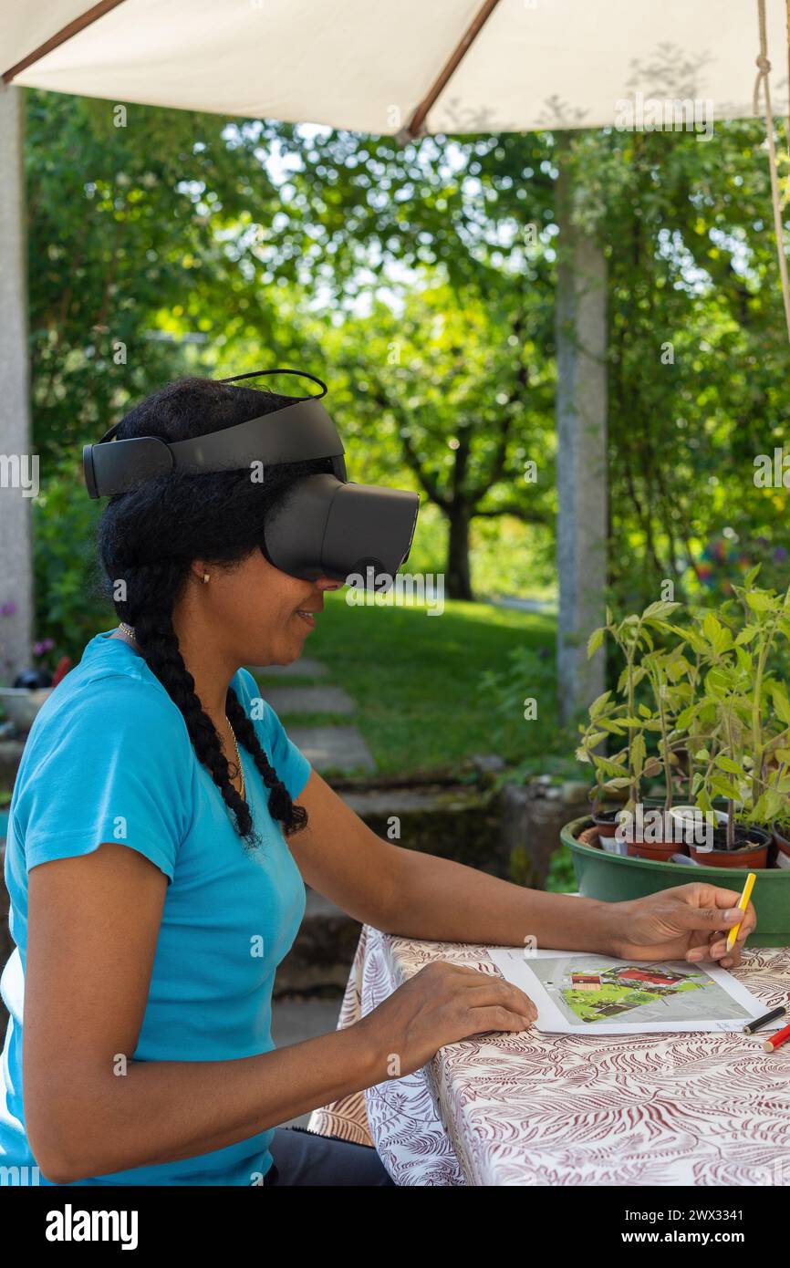 Une femme mixte dans des lunettes VR conçoit un plan de jardin avec des crayons de couleur à une table. Banque D'Images