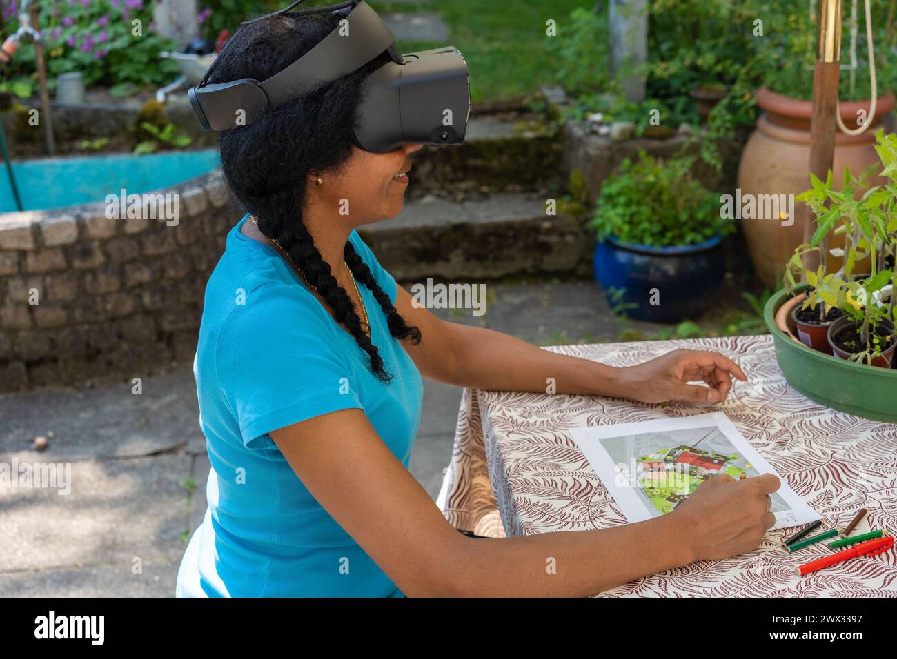 Une femme mixte portant des lunettes de réalité virtuelle est assise à une table de jardin, élaborant un plan de jardin. Banque D'Images