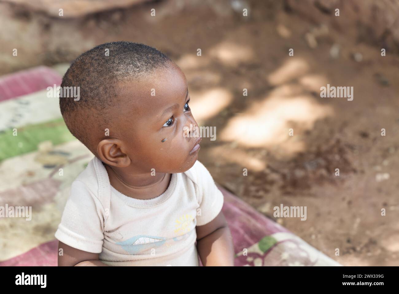 bambin africain célibataire, avec une mouche sur le visage, assis dehors sur une couverture devant la maison dans un village, Banque D'Images