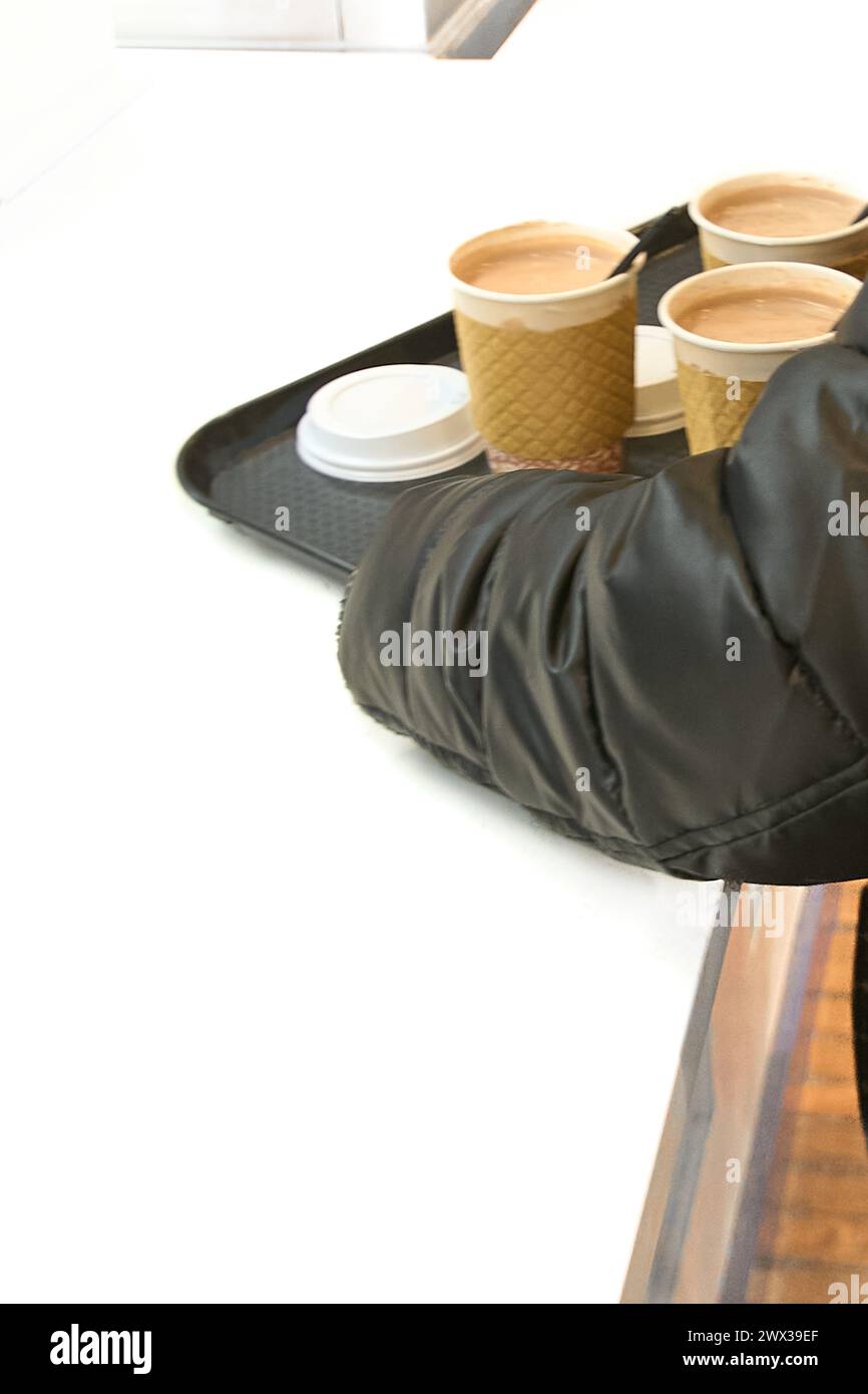 Un bras vêtu d'une veste noire tient un plateau avec quatre tasses de café à emporter, montrant un service rapide et efficace. Banque D'Images