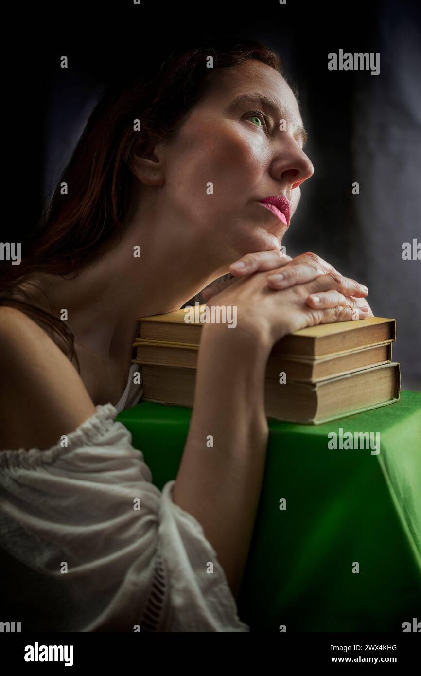 Femme dans une blouse blanche avec un décolleté Carmen avec ses mains reposant sur un vieux livre dans une attitude romantique dans le style de la prière, œuvre de Domeni Banque D'Images