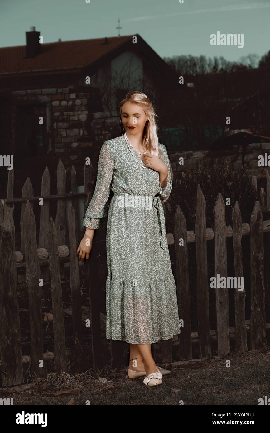 Une jeune femme aux cheveux blonds et Un sourire de rêve se tient devant une vieille clôture en bois dans une robe d'été Airy. L'air de nuit est chaud et plein de Su Banque D'Images