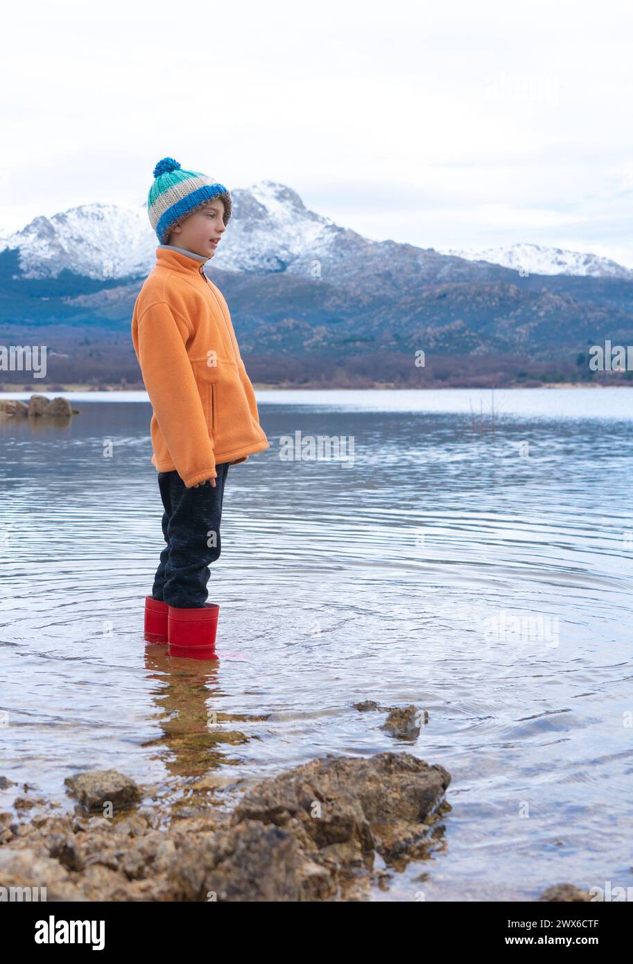 Garçon avec des bottes de pluie en hiver debout dans un lac avec des montagnes enneigées en arrière-plan Banque D'Images