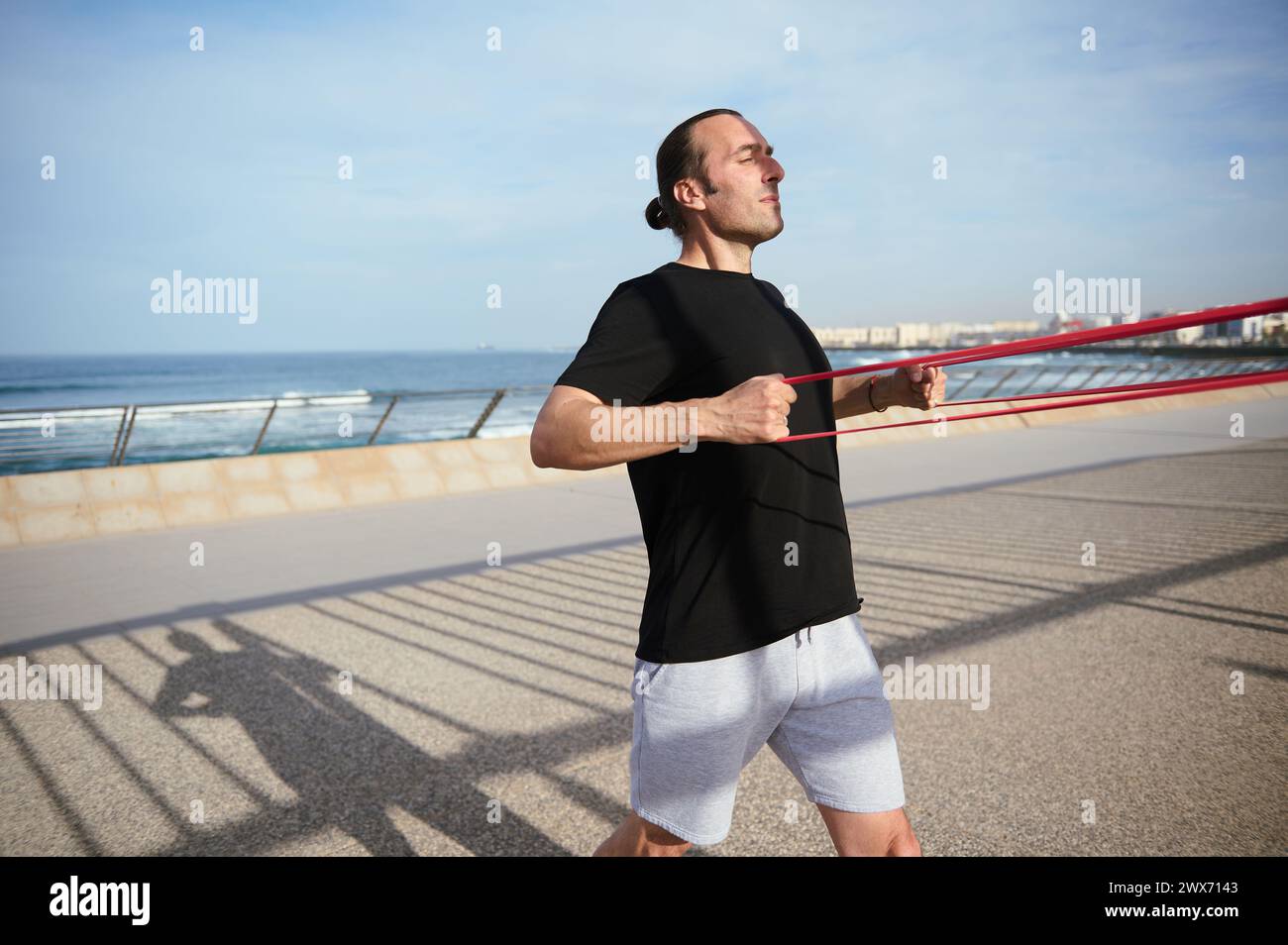 Homme sportif jeune adulte caucasien en t-shirt noir et short de sport gris, s'exerçant avec la bande de fitness de résistance sur la promenade de l'Atlantique, appréciant salut Banque D'Images