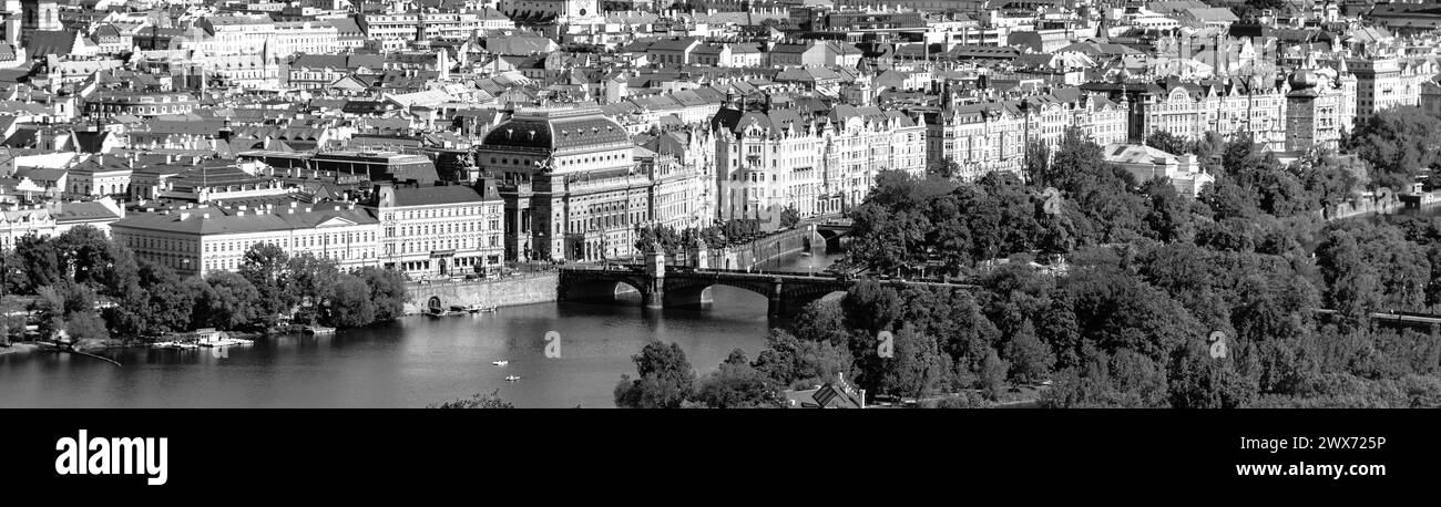 Une vue sur le Théâtre National de Prague, avec la rivière Vltava et un pont au premier plan entouré d'arbres et d'architecture. Prague, Tchéquie. Photographie noir et blanc. Banque D'Images