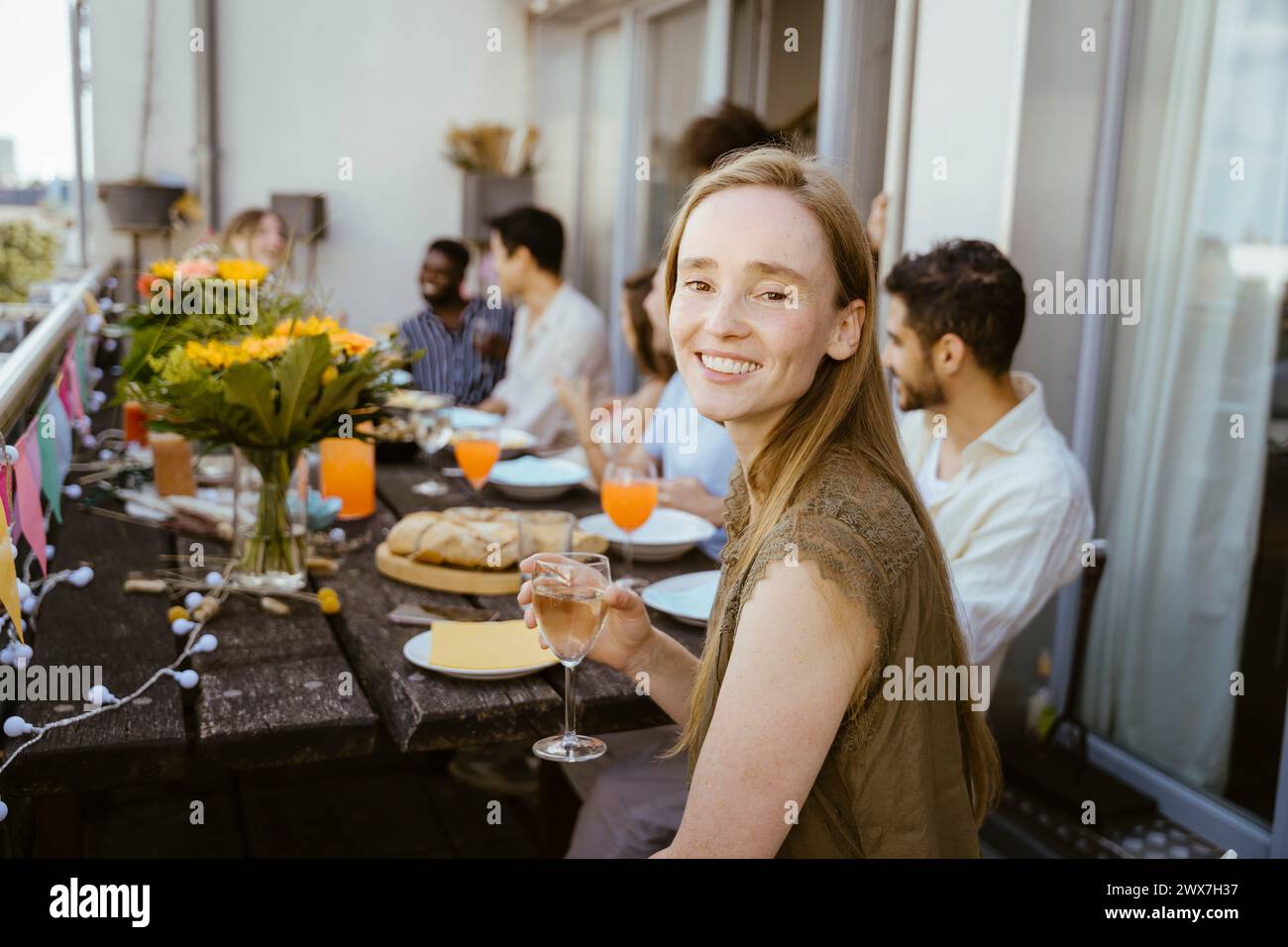 Vue latérale d'une femme souriante tenant du verre de vin tout en étant assise avec des amis à la table à manger pendant le dîner sur le balcon Banque D'Images