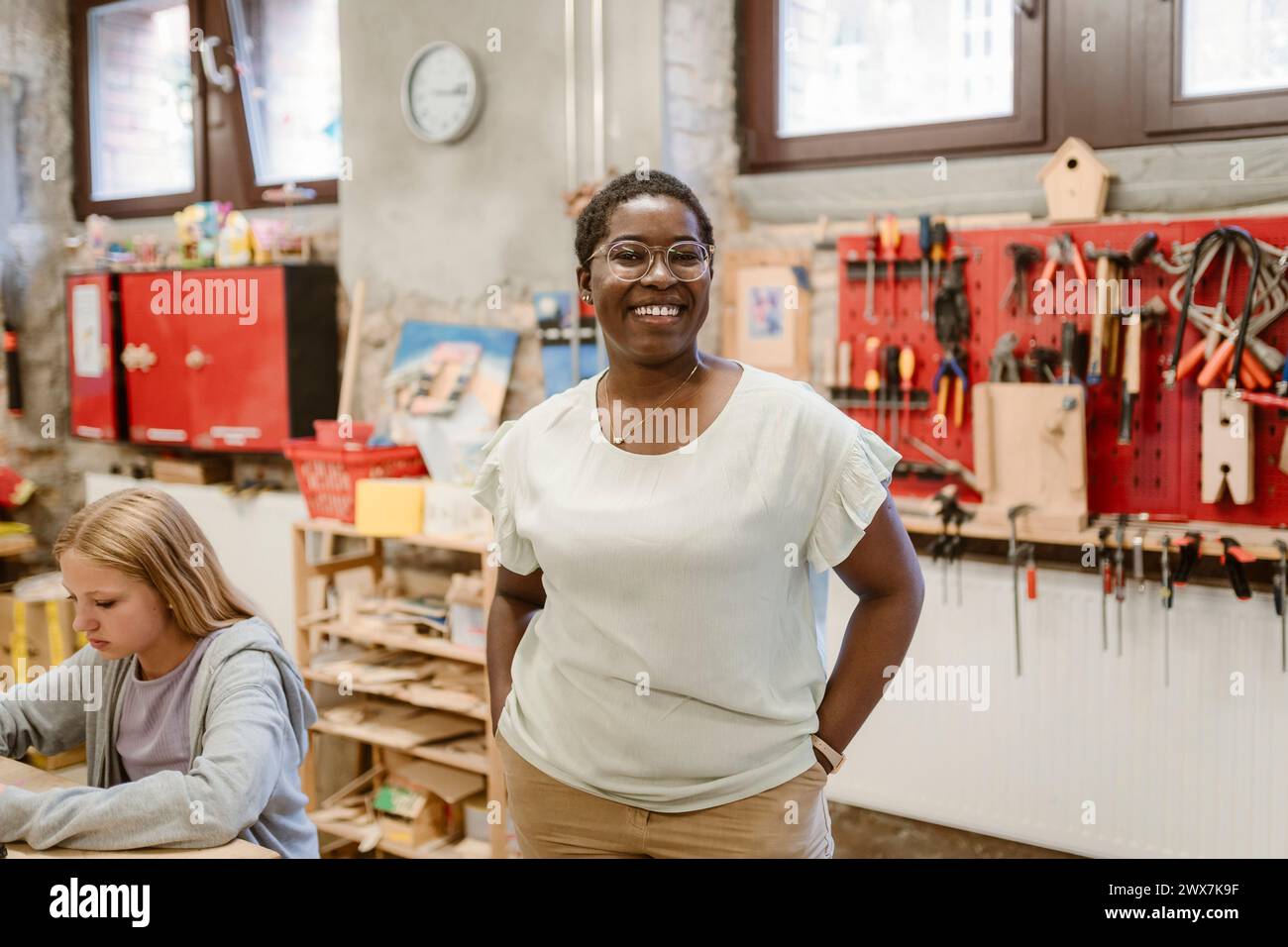 Portrait d'une enseignante heureuse debout avec les mains dans les poches à côté de la fille à l'atelier de l'école Banque D'Images