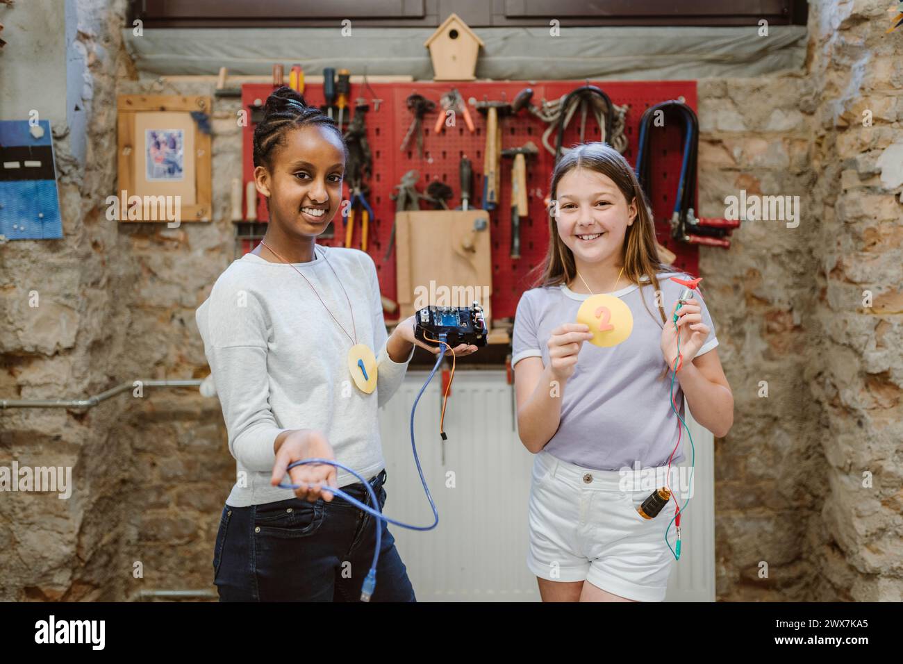 Portrait d'étudiantes souriantes montrant des réalisations et des pièces électriques tout en se tenant debout dans l'atelier de technologie à s. Banque D'Images