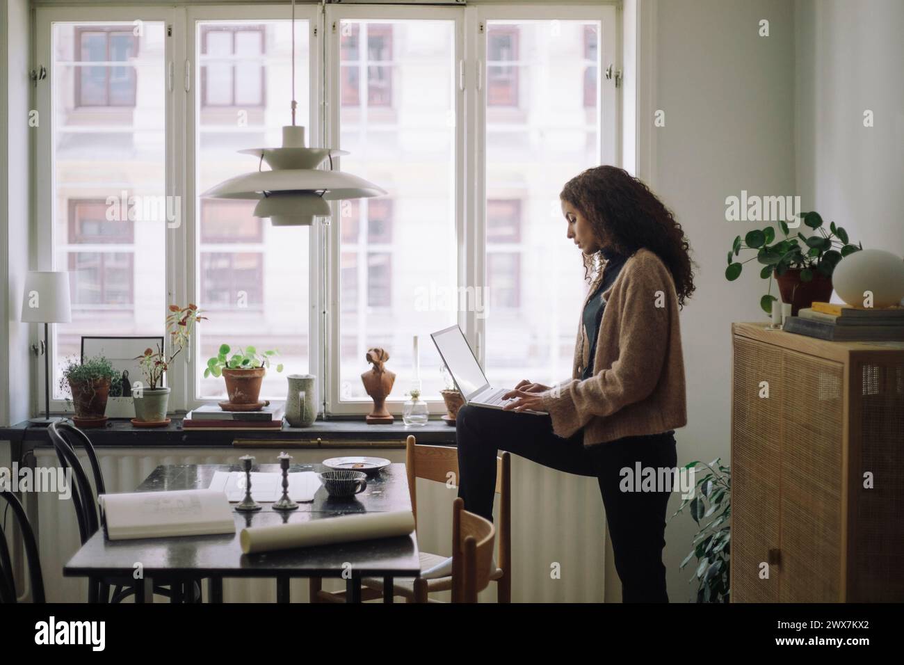 Vue latérale de l'ingénieur de construction féminin utilisant un ordinateur portable tout en se tenant debout avec une jambe sur la chaise près de la table à la maison Banque D'Images