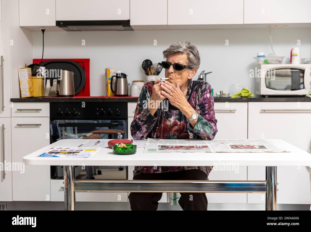 Une vieille dame avec des lunettes noires allume une cigarette dans la cuisine devant son journal et son cendrier. Banque D'Images