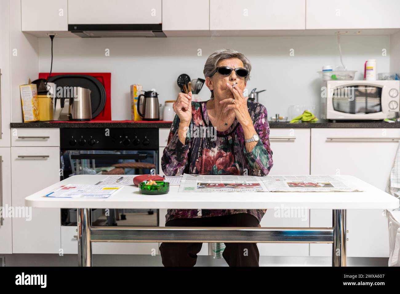 Une vieille dame pleine de rides fume confortablement à la maison dans sa cuisine portant des lunettes de soleil noires. Banque D'Images