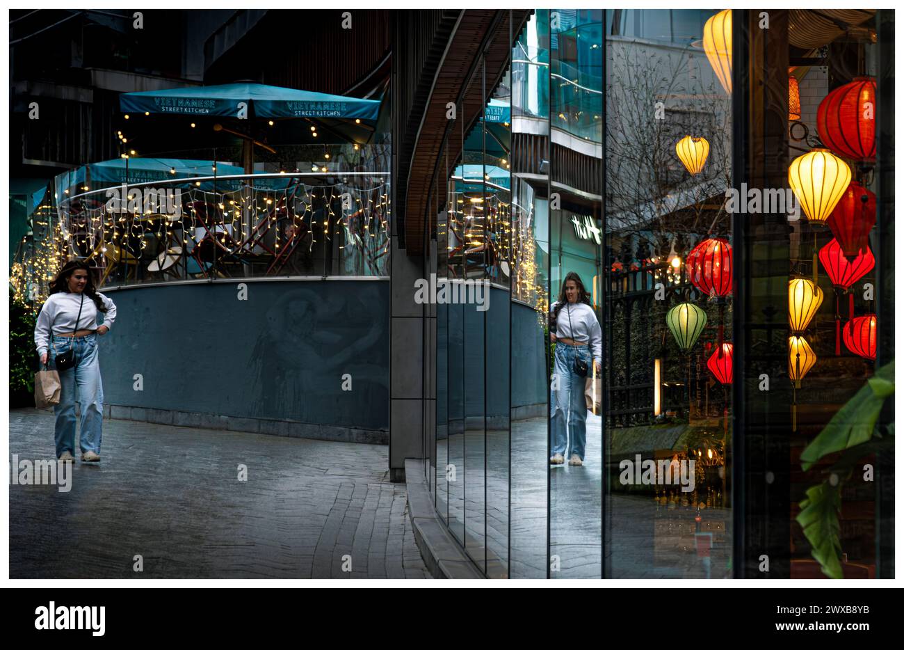 Jeune femme et ses environs se reflètent dans la fenêtre du restaurant du centre-ville. Le restaurant dispose de plusieurs lumières colorées et éclairées de style lanterne Banque D'Images