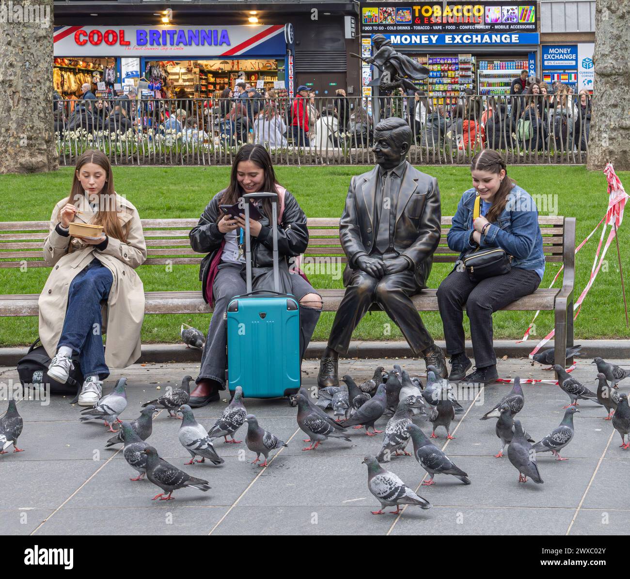 Femmes assises sur un banc à côté d'une statue de bronze de Mr Bean, à Leicester Square, Londres. Les pigeons se rassemblent devant eux à la recherche de nourriture. Banque D'Images