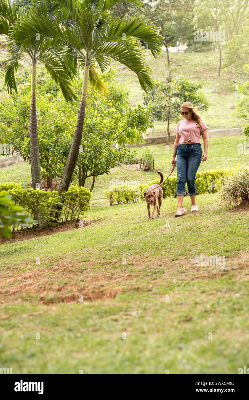 Femme avec des lunettes de soleil marchant aux côtés de son chien, son fidèle compagnon, dans le parc par une journée ensoleillée. Banque D'Images