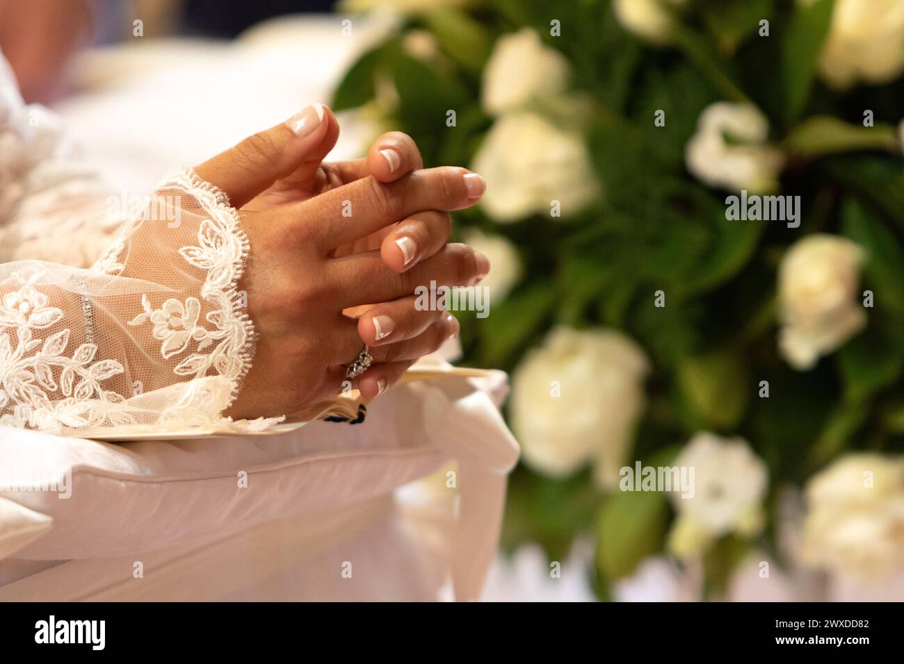 les mains d'une mariée, ornées d'une bague de mariage délicate, sont pliées dans la prière sur sa robe de dentelle détaillée, avec des roses blanches en arrière-plan Banque D'Images