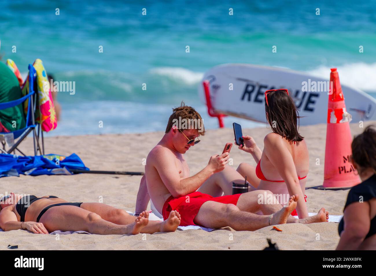 Fort Lauderdale, FL, États-Unis - 30 mars 2024 : jeune couple ensemble sur la plage mais vivant séparé vit sur ses téléphones Banque D'Images