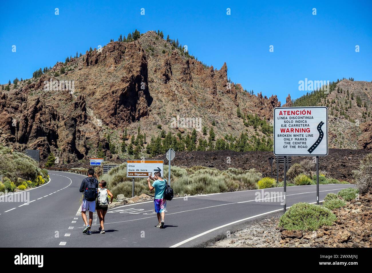 Groupe de randonneurs marchant le long de la route TF-21 dans le parc national du Teide avec des panneaux d'avertissement dans un paysage montagneux sous un ciel bleu clair Banque D'Images