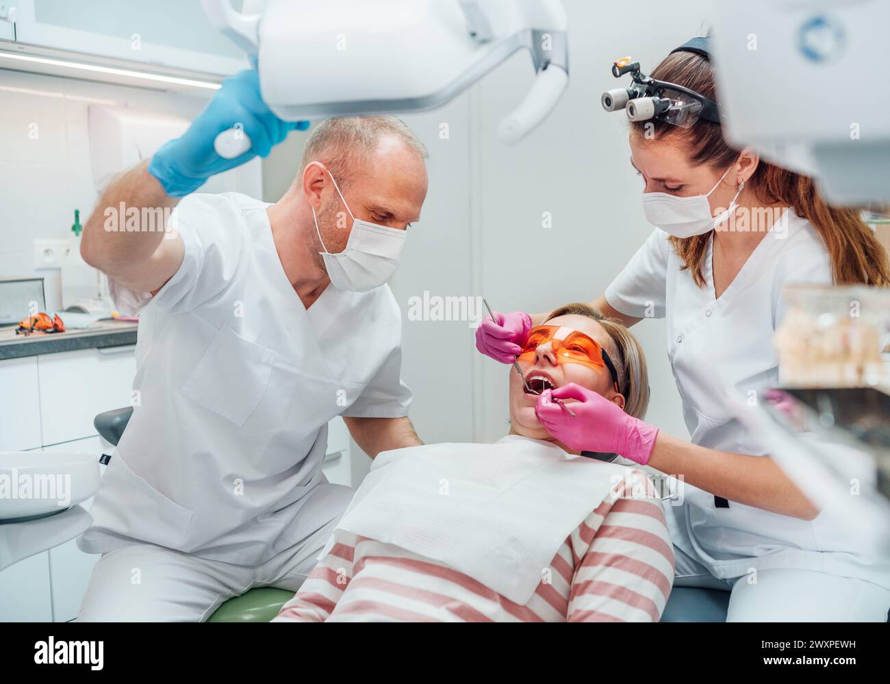 Rendez-vous en clinique dentaire. Femme dentiste en loupe faisant la prévention des dents à l'aide d'outils médicaux. Jeune homme assistant pointant la lumière. Banque D'Images
